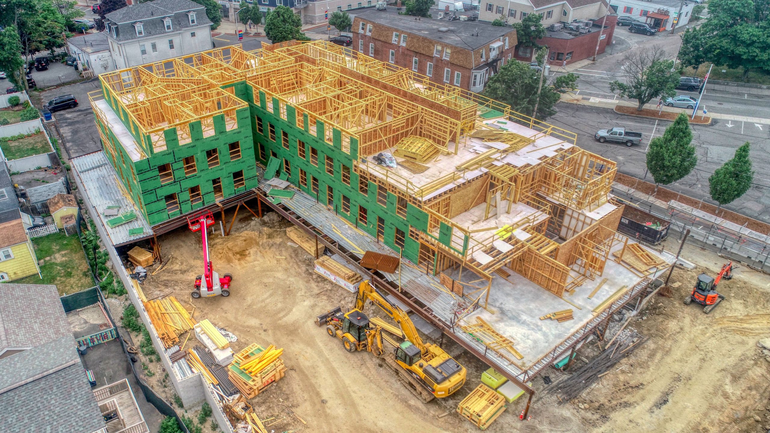Aerial view of a construction site with a green-framed building, yellow wood scaffolding, and machinery. Surrounding residential area visible.