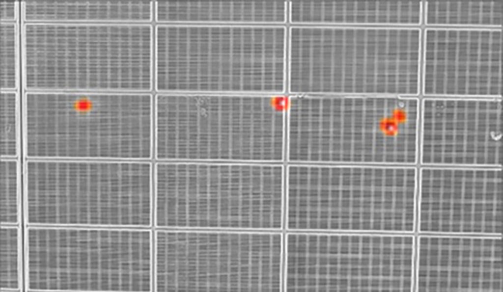 A thermal image of solar panels shows several bright orange hot spots against a gray grid pattern, indicating heat anomalies.