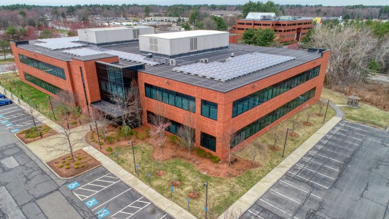 Aerial view of a red brick office building with solar panels on the roof, surrounded by empty parking lots and trees under an overcast sky.
