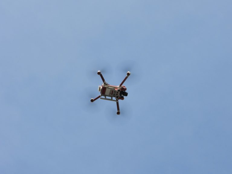A drone with four propellers flies against a clear blue sky, captured from below. The scene conveys innovation and technology in a serene setting.