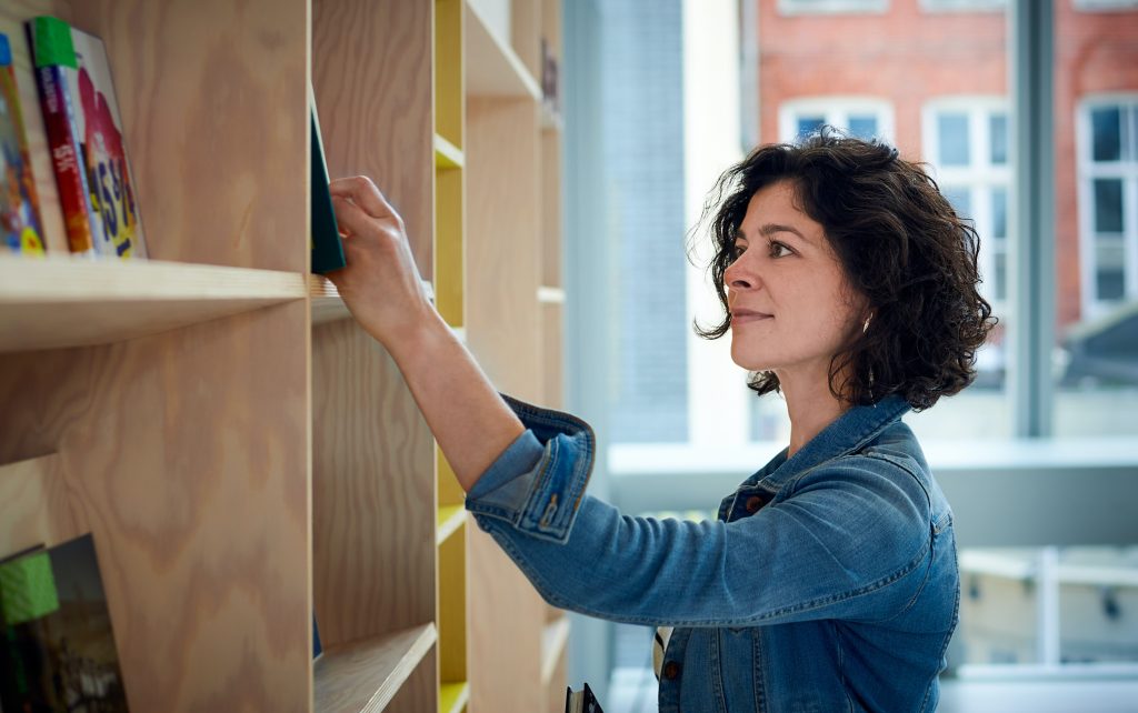 Woman in denim jacket reaching for a book on a wooden shelf in a bright room with large windows. Colorful books partially visible.