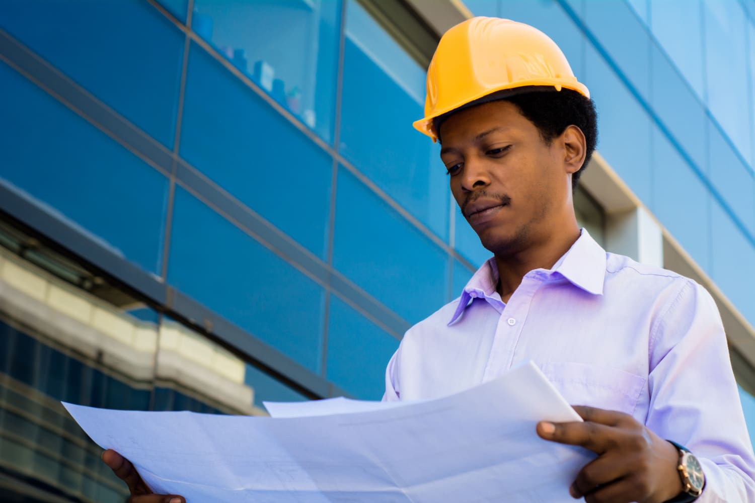 A focused engineer wearing a yellow hard hat examines blueprints outside a modern glass building, conveying professionalism and concentration.