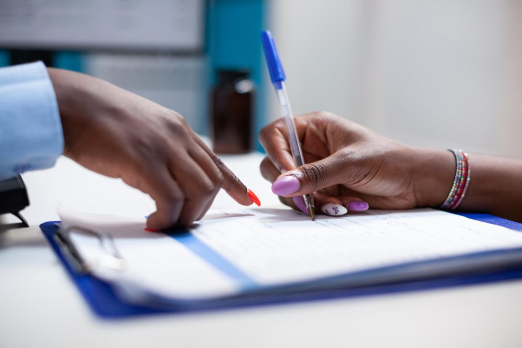 Close-up of two hands over a clipboard; one hand is holding a pen, poised to write, while the other points at a document, suggesting guidance or instruction.