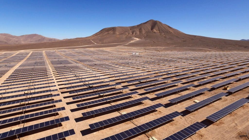 Expansive solar farm in a desert, with rows of solar panels under a clear blue sky and a mountain in the background, conveying sustainability.