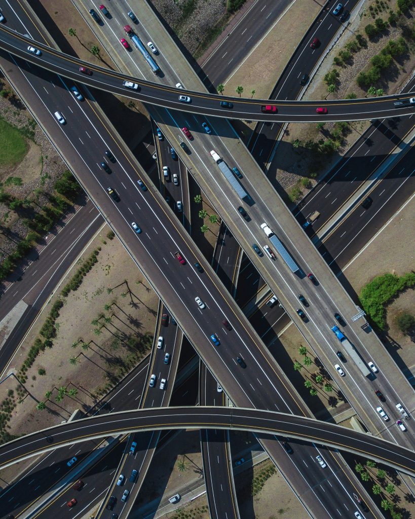 Aerial view of a complex highway interchange with multiple overpasses and roads crossing. Vehicles in various colors navigate the lanes. Sparse greenery and palm trees dot the landscape.