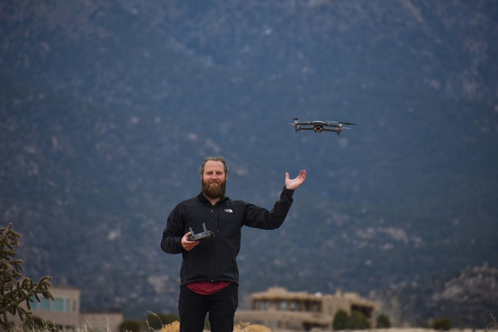 A man with a beard stands in a rugged landscape, flying a drone with a remote control. The sky is overcast, and mountains are in the background.