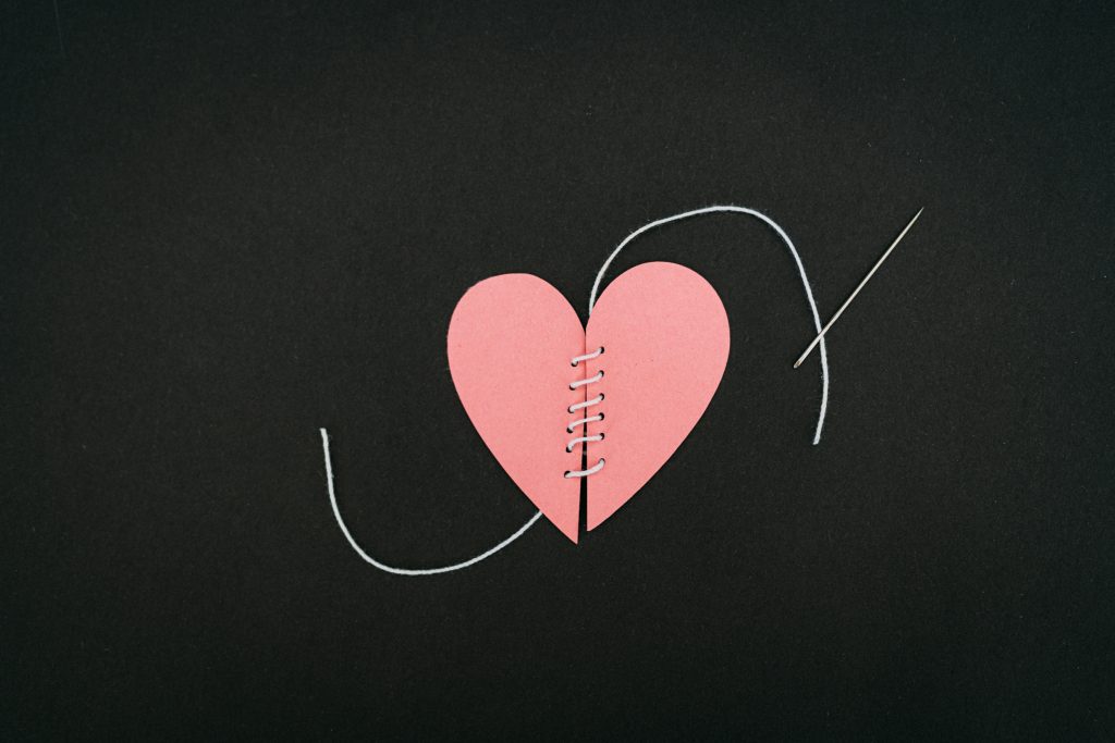 A pink paper heart sewn together with a black thread and needle on a dark background.