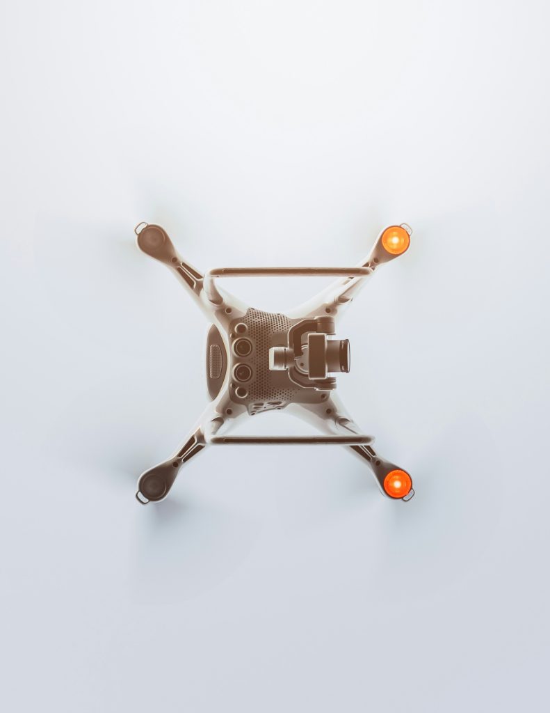 A drone viewed from above, with a camera and two glowing orange lights on its arms. The background is a soft, light gray, creating a sleek, modern tone.