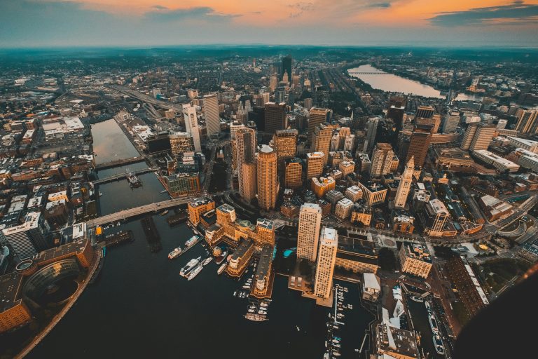 Aerial view of a cityscape at sunset, with tall buildings, a busy river with boats, and a glowing sky, conveying an urban, vibrant atmosphere.