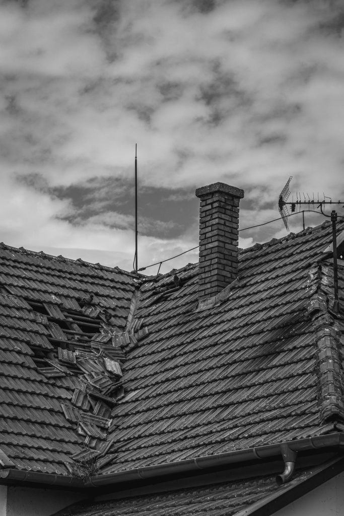 A damaged roof with broken tiles and a visible hole, next to a brick chimney. An old TV antenna is attached. The sky is cloudy, creating a somber mood.