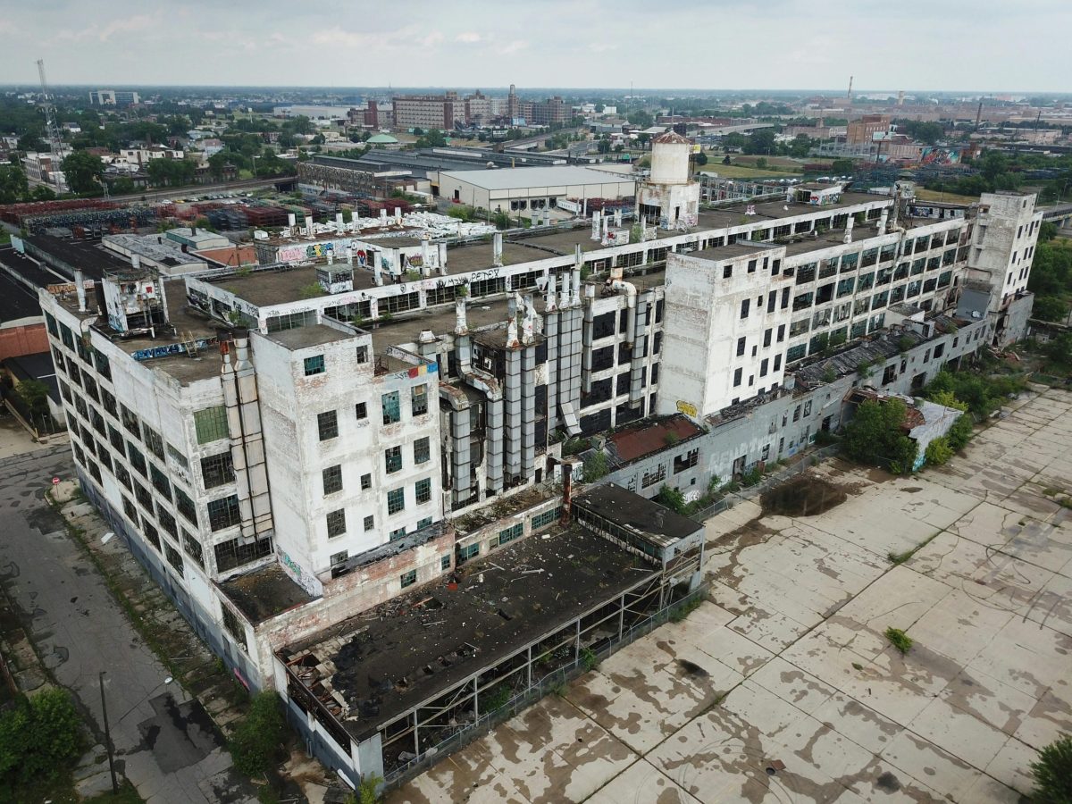 Aerial view of a large, abandoned industrial building with peeling white paint and broken windows. Surrounding area is overgrown and desolate, conveying neglect.