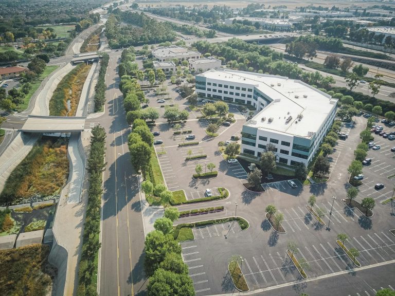 Aerial view of a large office building with a white roof shaped like a right angle, surrounded by several parking lots with scattered cars and lush green trees, adjacent to a river flowing under bridges. The scene conveys a sense of spaciousness and order.
