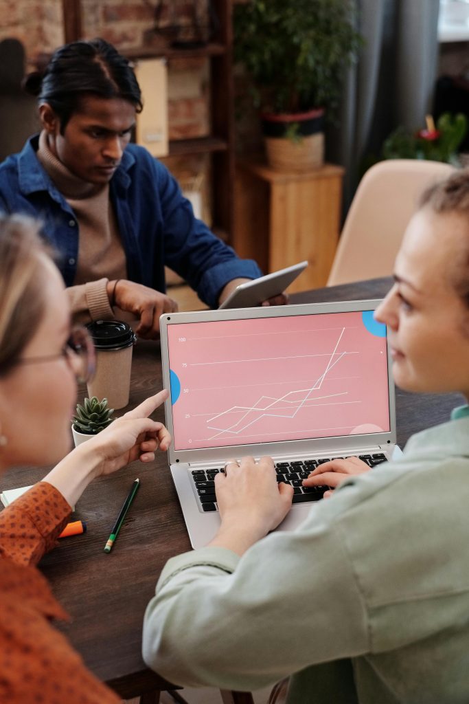 Two women discuss a graph on a laptop screen, pointing with focus. A man in the background reads a tablet. The setting is a casual office.