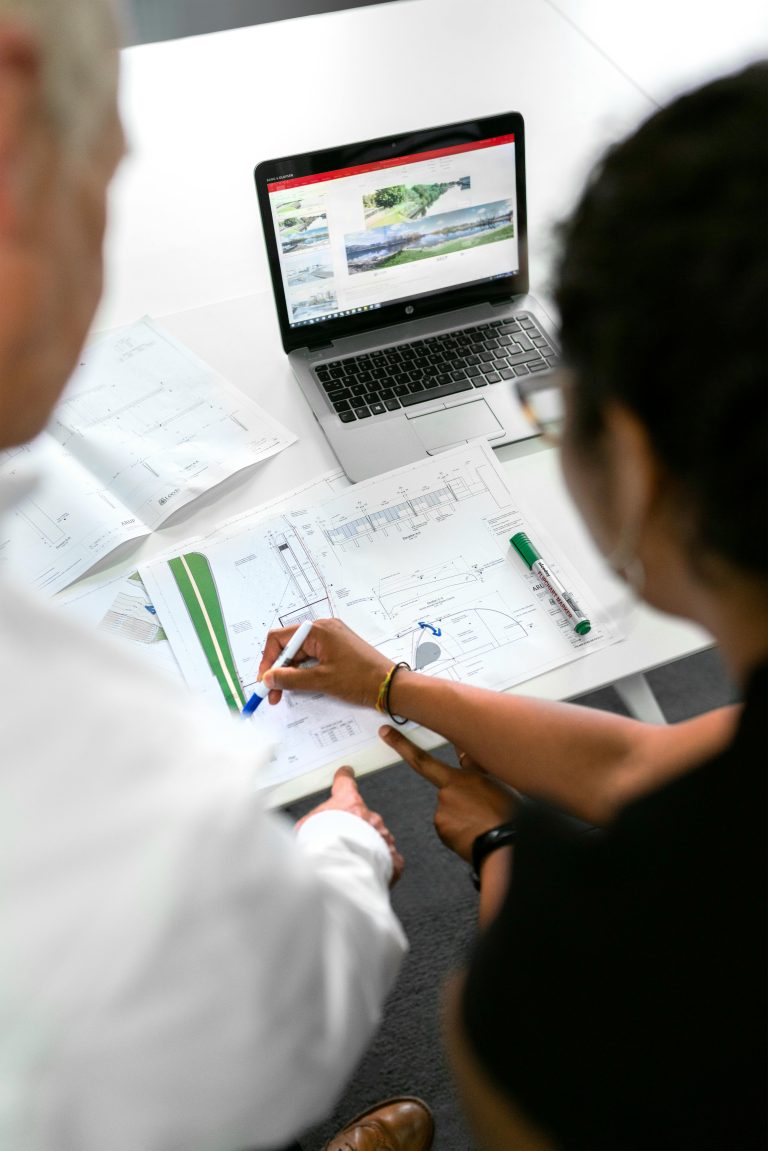 Two people collaborate on architectural plans at a desk. A laptop displaying images is open, and markers are scattered on the plans.