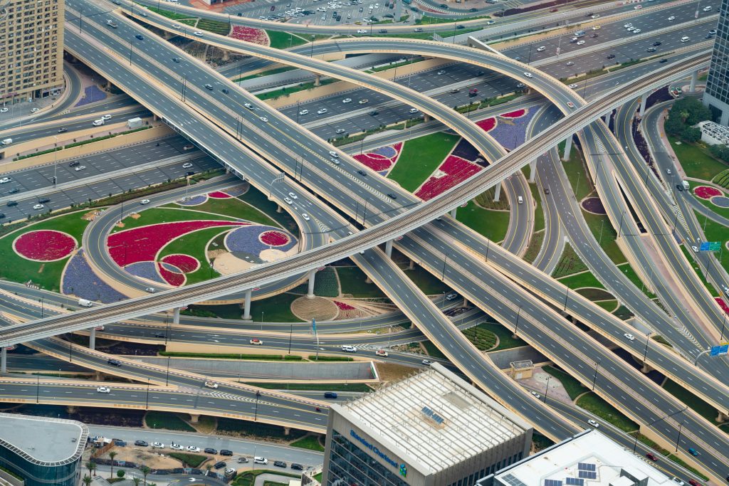 Aerial view of a complex highway interchange with multiple overlapping roads. Surrounding green spaces feature red and purple geometric landscaping.