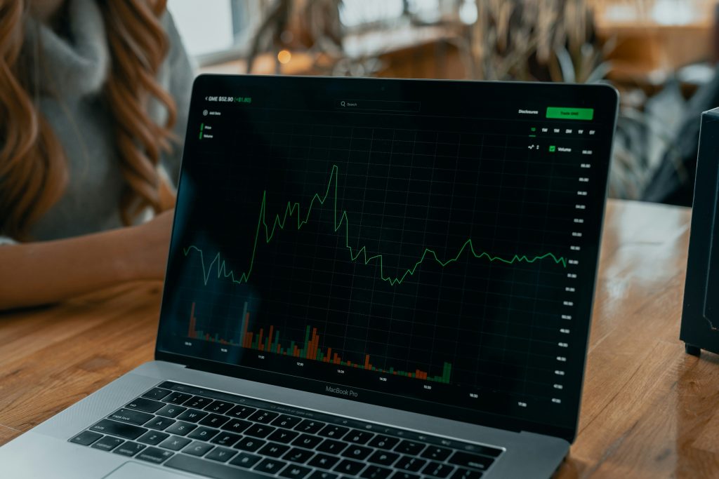 A laptop on a wooden table displays a green line graph with bar chart data, suggesting stock or financial analysis. The setting is informal and relaxed.